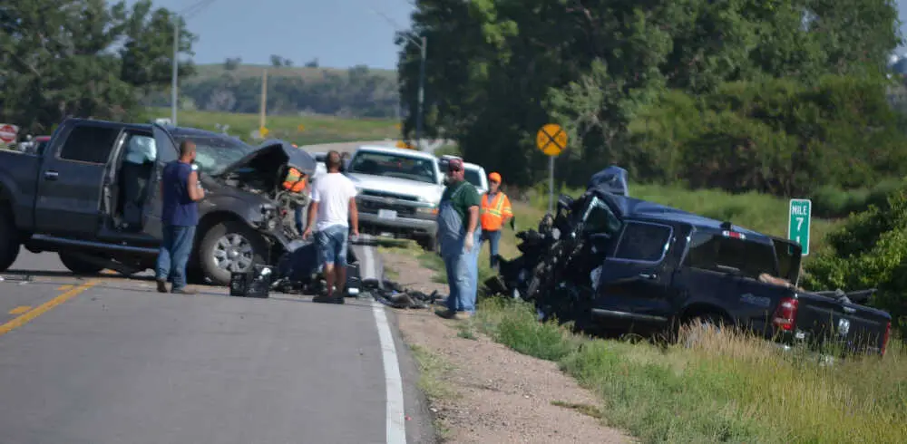 Two heavily damaged pickup trucks after a head-on collision on a rural road, with several people and emergency workers at the scene