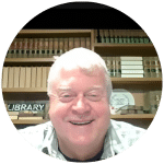 An older man with short white hair, wearing a light plaid shirt, smiles at the camera while sitting in front of bookshelves filled with books and files.