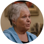 An older woman with short gray hair, wearing a light blue jacket over a black top, looks to the side while speaking indoors.